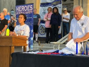 Ingrid Ortega stands at a podium delivering a speech during the Beyond the Badge 5K, with Jerry Baker at her side.