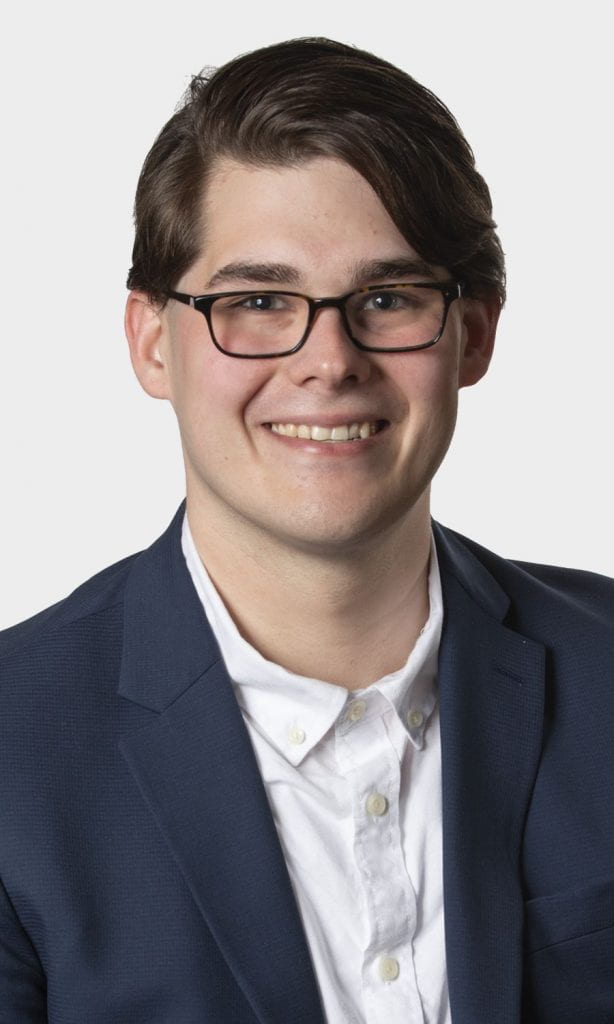 Kyle Casteel smiles, wearing a white button-up shirt, blue blazer and dark glasses. 