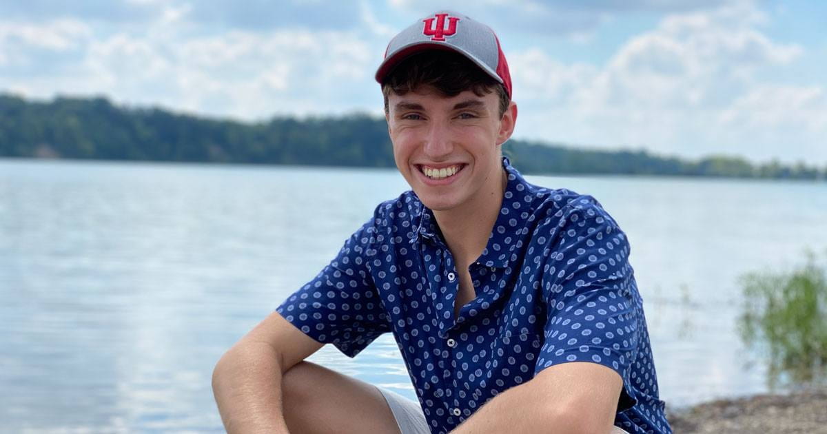 man sits in front of lake with IU hat on and blue button-up shirt