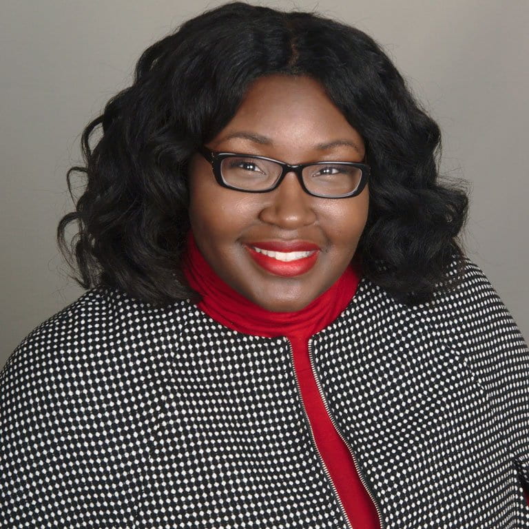 headshot of woman wearing glasses, red shirt, and black/white check jacket