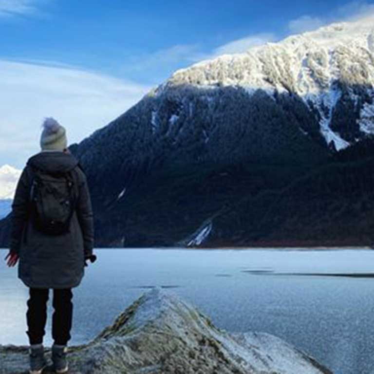 woman stands with back to camera facing snowcapped mountain by frozen lake