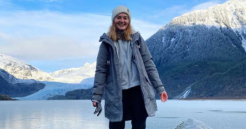 woman stands in front of snowcapped mountain by frozen lake
