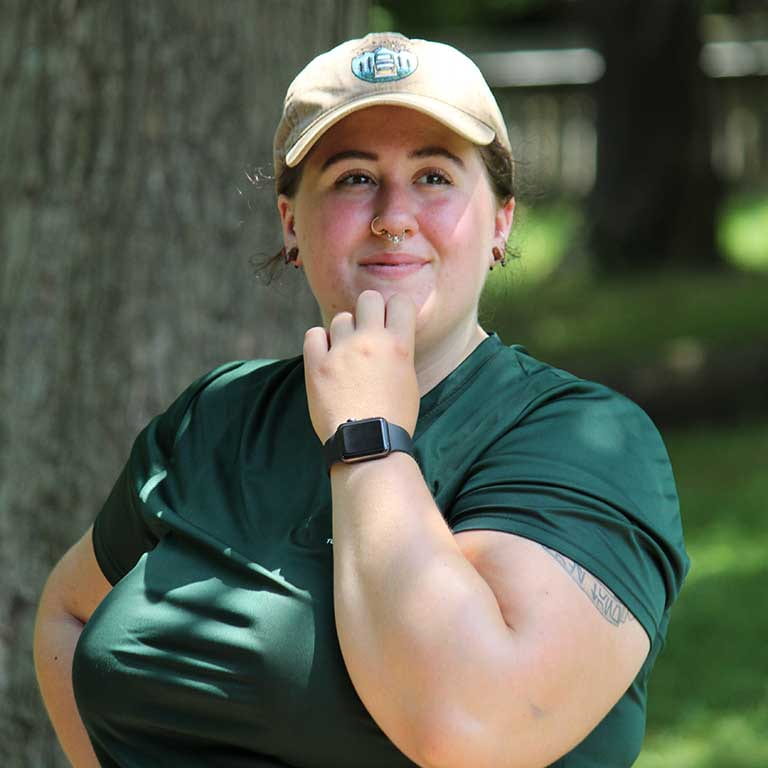 Photo of Quinlin Malloy in a green shirt and khaki hat with her hand on her chin and trees behind her.