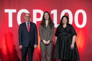 Three people in business attire stand in front of a red curtain with a Top 100 sign behind them.