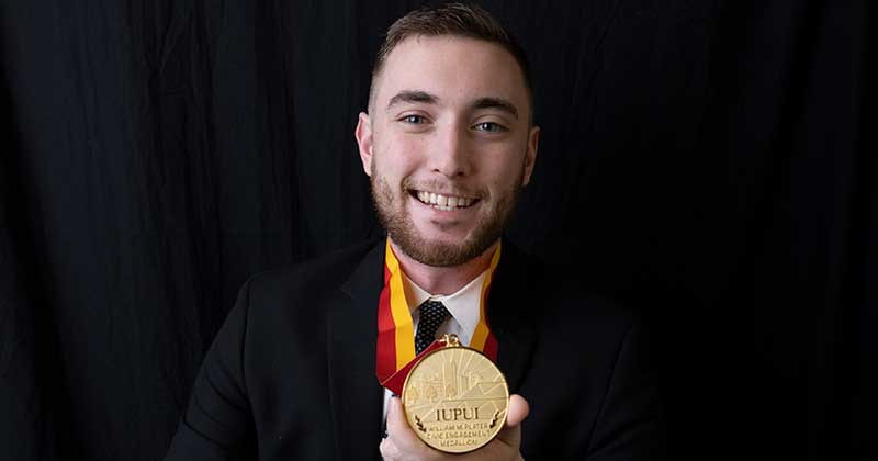 Photo of man with beard holding medal, wearing a black suit jacket and smiling.