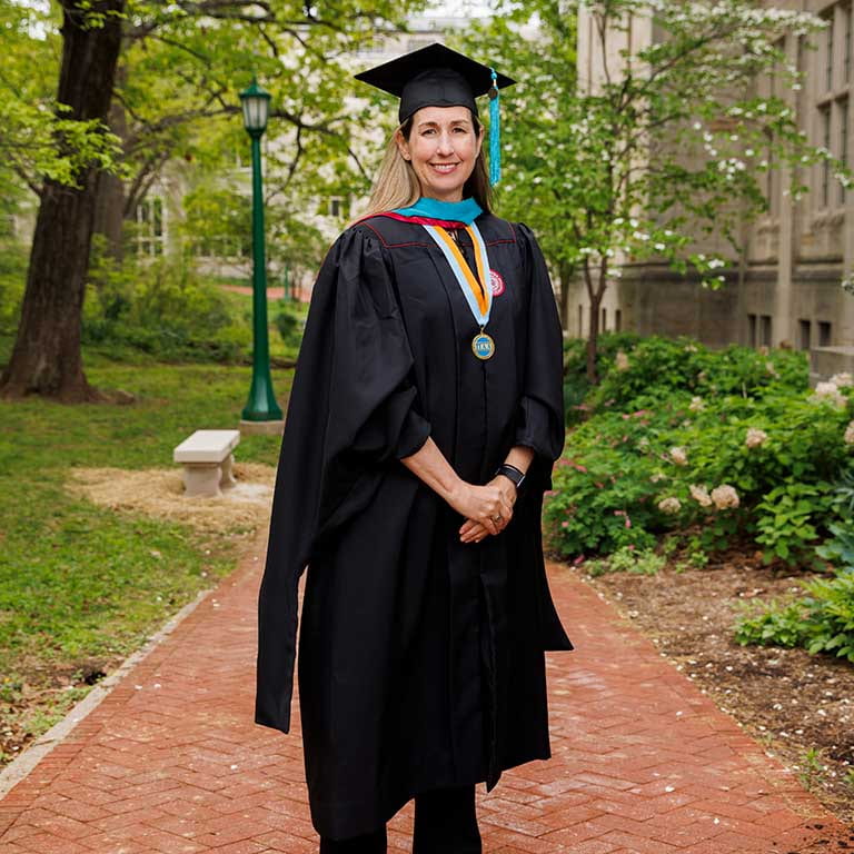 Woman stands in graduation cap and gown on a brick sidewalk with green trees behind her.
