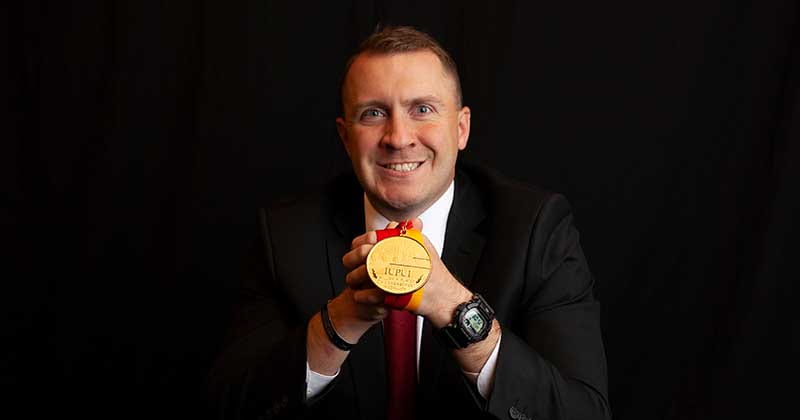 Man in a suit holds a gold medal in front of him while smiling.