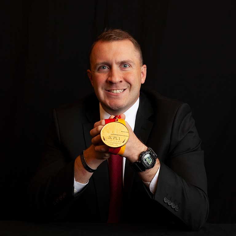 Man in a suit holds a gold medal in front of him while smiling.