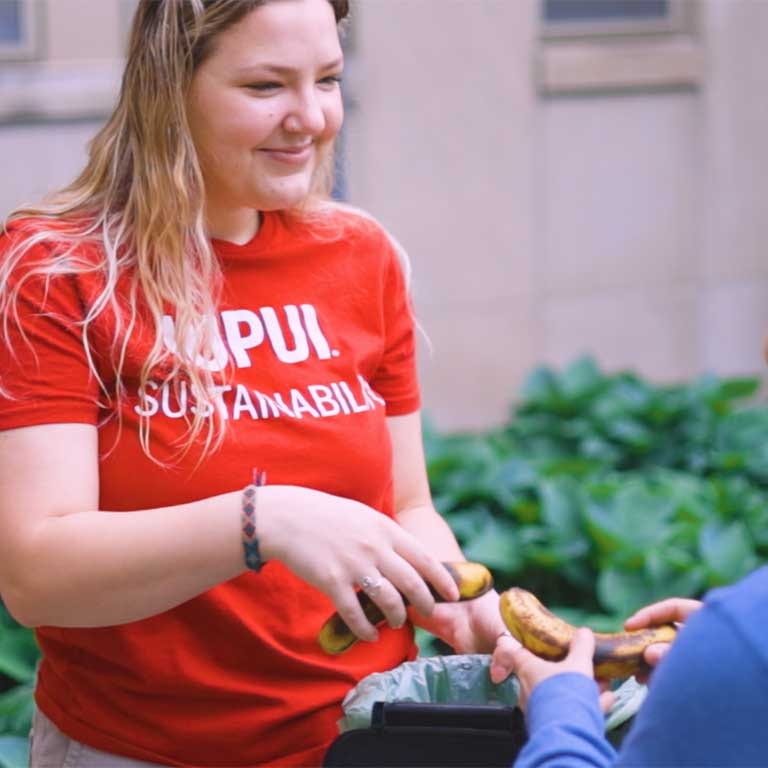 Woman in red IUPUI Sustainability shirts holds a composting bin as she helps another students put bananas in the bin.