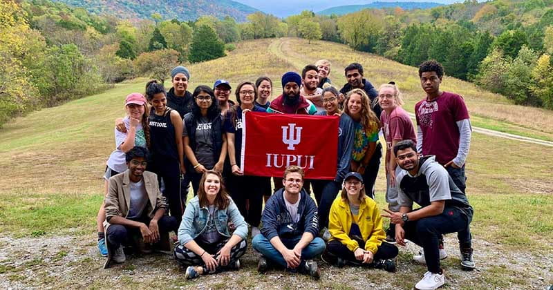 Group of students stand in a field with the mountains behind them, holding an IUPUI flag.
