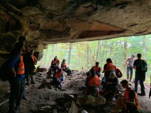 A group of students sits under the opening of a cave wearing orange vests and hard hats as they learn about ecosystems.
