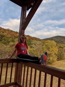 woman sits on porch rail with mountains behind her with fall leaves on the trees.