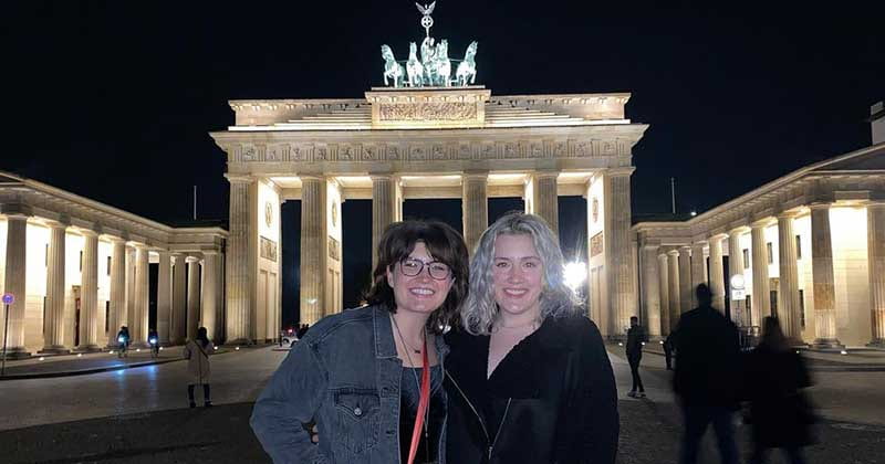 Two women stand in front of a monument in Berlin at night. The monument is brightly lit in the background.