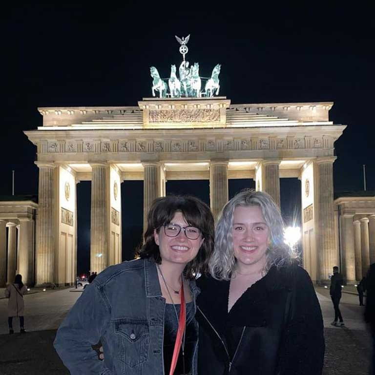 Two women stand in front of a monument in Berlin at night. The monument is brightly lit in the background.