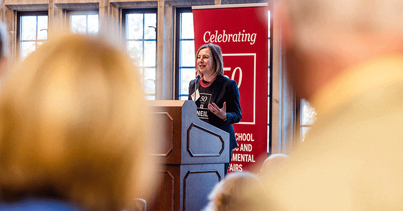 A woman stands at a wood podium, speaking to a crowd. A tall red banner is behind her with the words celebrating 50 years. In the foreground, we see two people watching the woman speak.