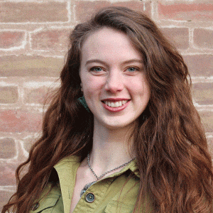 A woman with long brown hair stands against a brick wall, smiling while wearing a green button-up shirt
