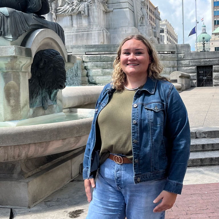 Woman stands in denim jacket and green shirt, smiling outside the Soldier and Sailors Monument in Indianapolis.