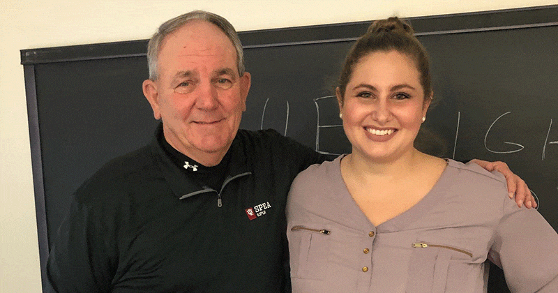 Faculty member wearing a black O'Neill shirt stands with his arm around a former student, wearing a mauve shirt. They're in a classroom with a chalkboard behind them.