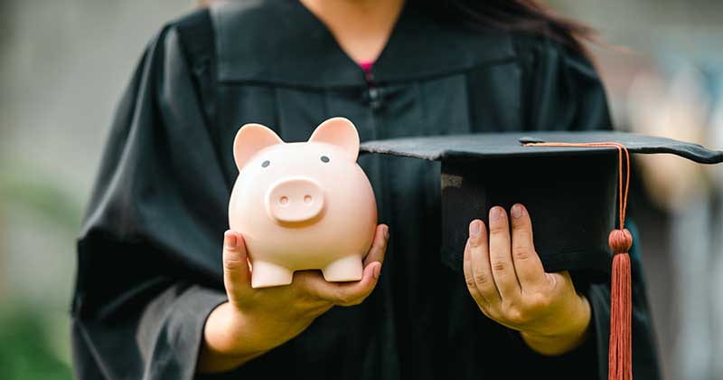Graduating student in their cap and gown hold a piggy bank in one hand, and their graduation cap in the other.