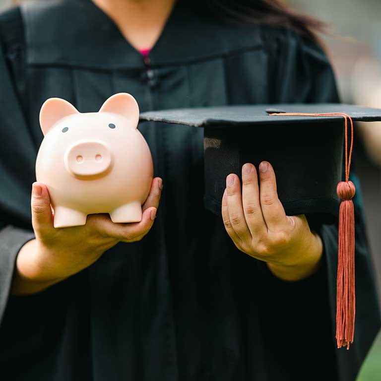 Graduating student in their cap and gown hold a piggy bank in one hand, and their graduation cap in the other.