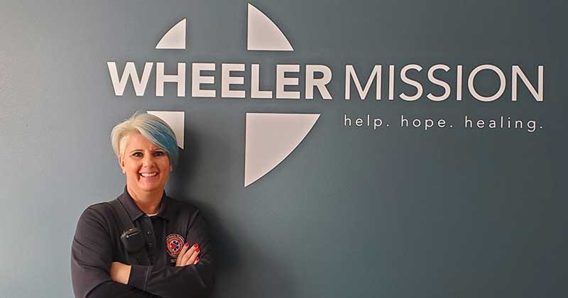A smiling woman stands in front of a wall with a Wheeler Mission logo on it, with her arms crossed.