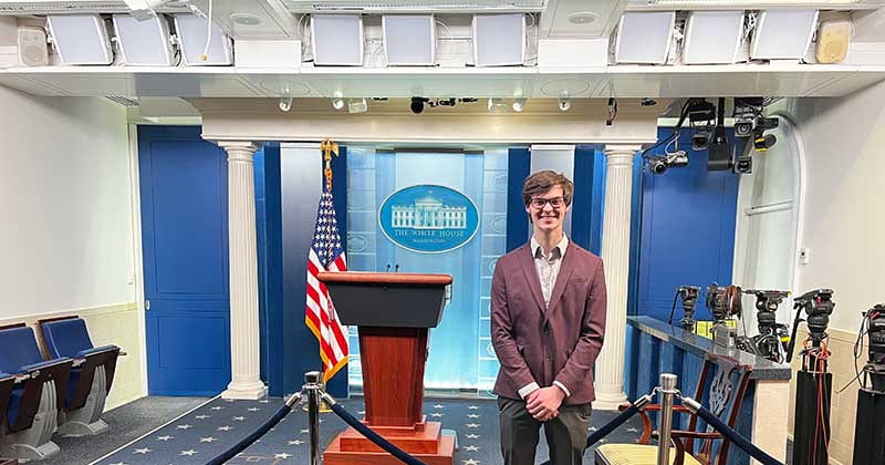 Image of the White House Press briefing room with a man in a suit jacket standing in front of the podium, smiling.