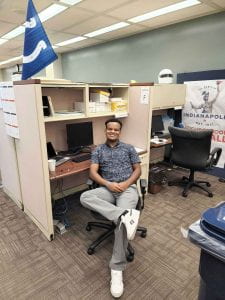 A man sitting in a chair at a desk in his office smiles. There's a Colts flag next to him and a banner for the city of Indianapolis.