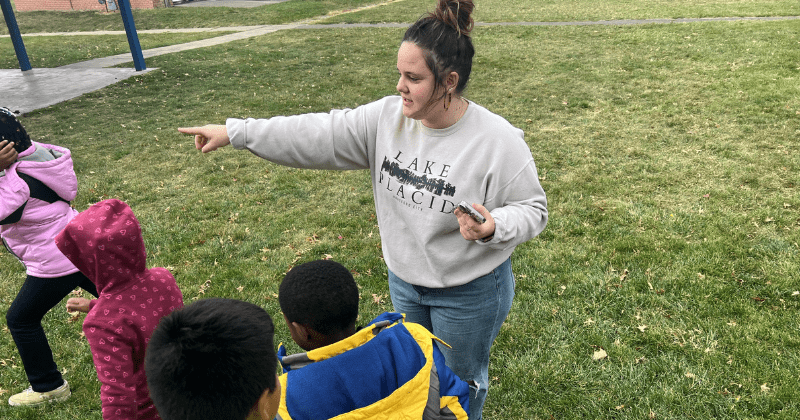 A woman stands in the grass directing a group of young children wearing coats to line up.