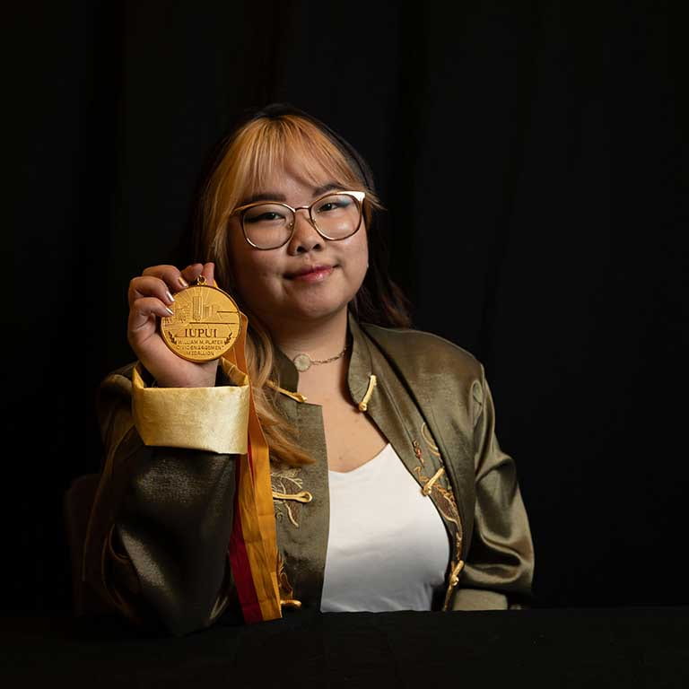 A woman sits at a table with a black background behind her. She's wearing a green jacket and holding a medallion.