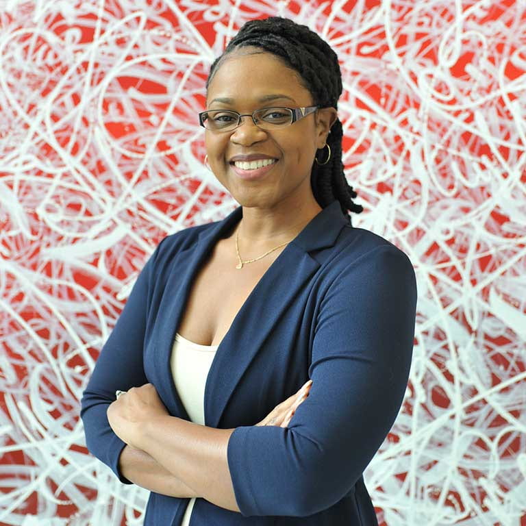 A woman wearing glasses stands in front of a red and white background, smiling. She's wearing a navy blue blazer and a white shirt.