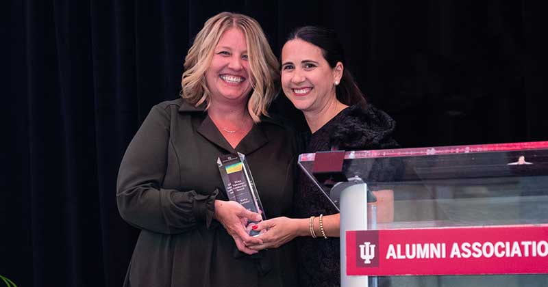 Two women stand on stage smiling while one presents the other with an award.