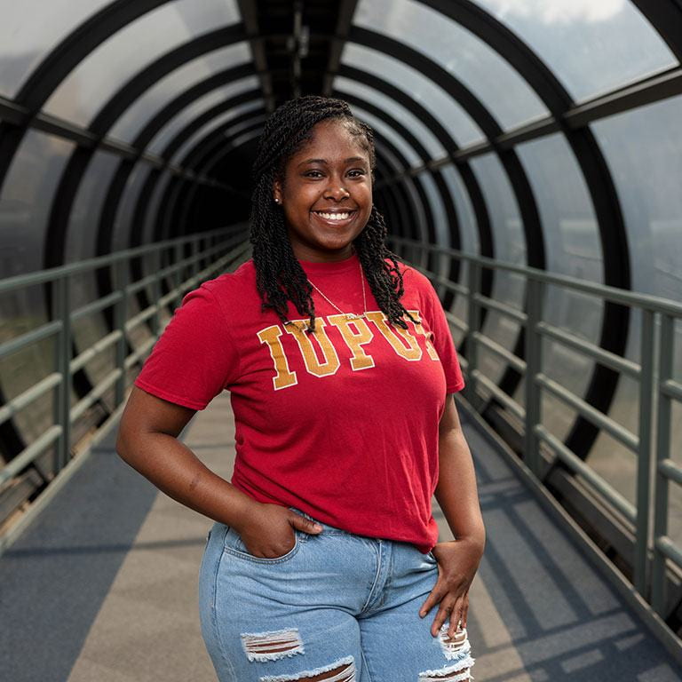 A woman stands in a pedestrian tube wearing a red IUPUI shirt. She's smiling and has both hands in the pockets of her jeans.