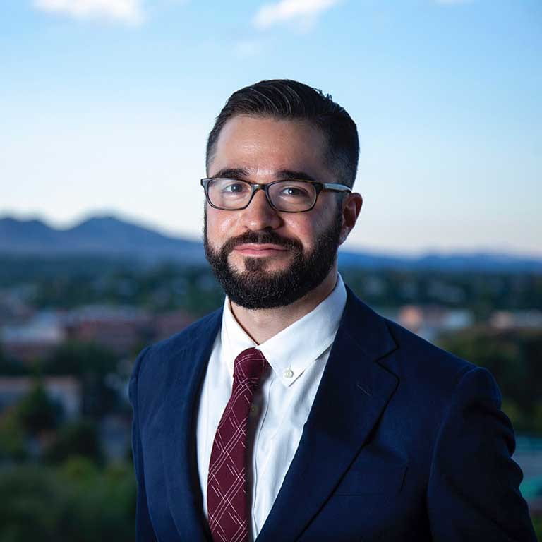 A man with a beard and glasses is wearing a suit. In the background is a city and a mountain skyline.