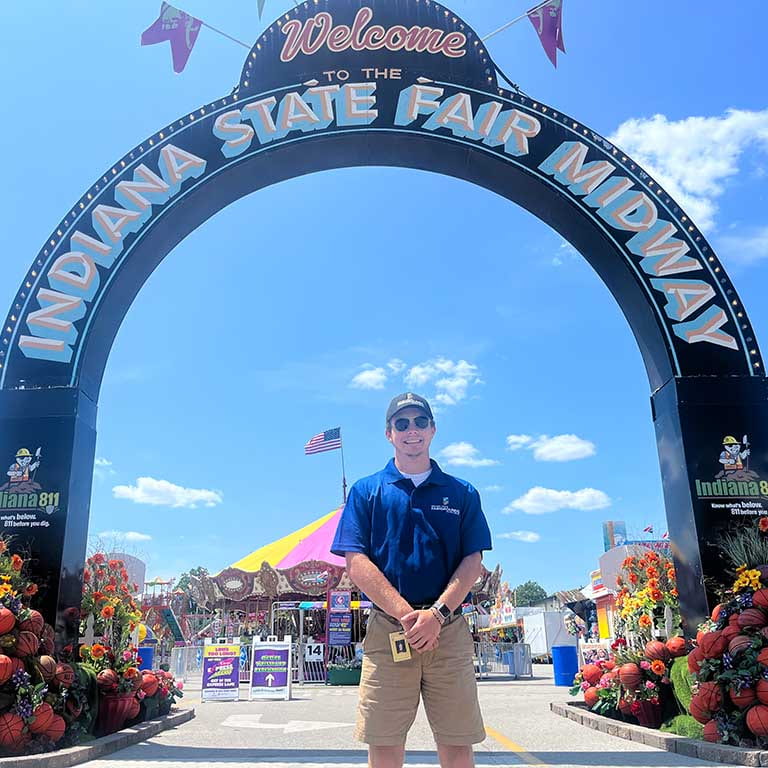 A man stands outside in front of the midway sign at the Indiana State Fair. The sky is blue with a few clouds. He's wearing a public safety uniform.