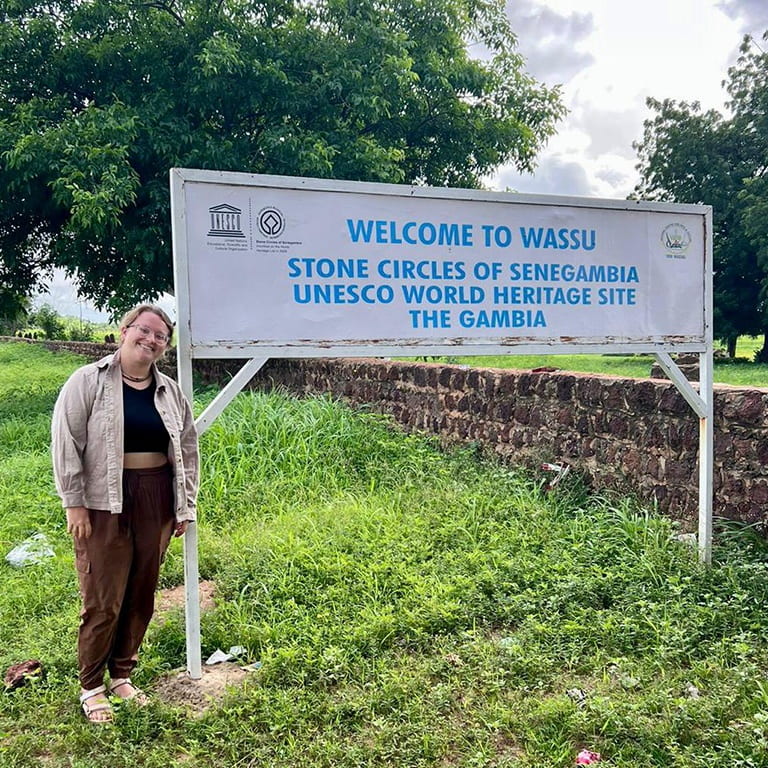 A woman stands next to a sign that reads Welcome to Wassu.