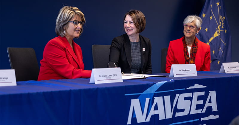 Three women sit at a table at a news conference to sign documents.