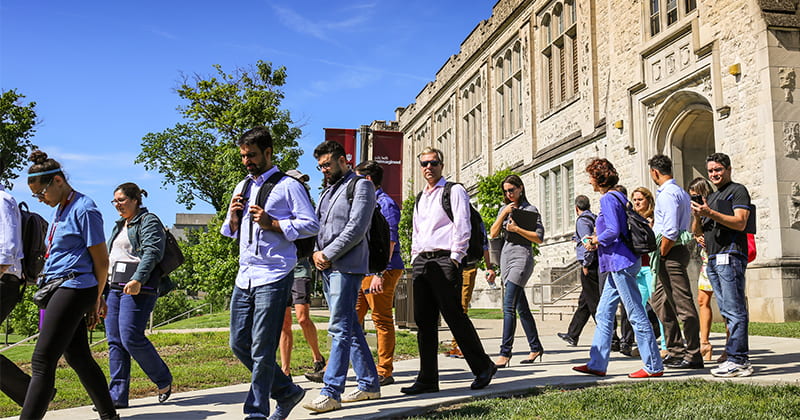 A group of people walk outside on a sidewalk on a bright sunny day with a university building behind them.