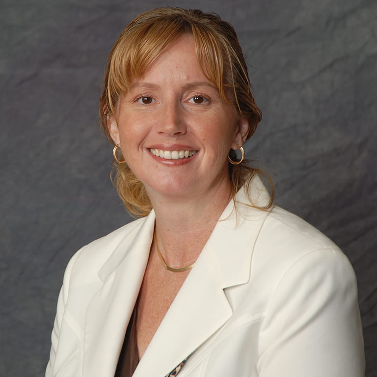 A woman with light reddish hair sits smiling in front of a gray backdrop while wearing a white blazer.